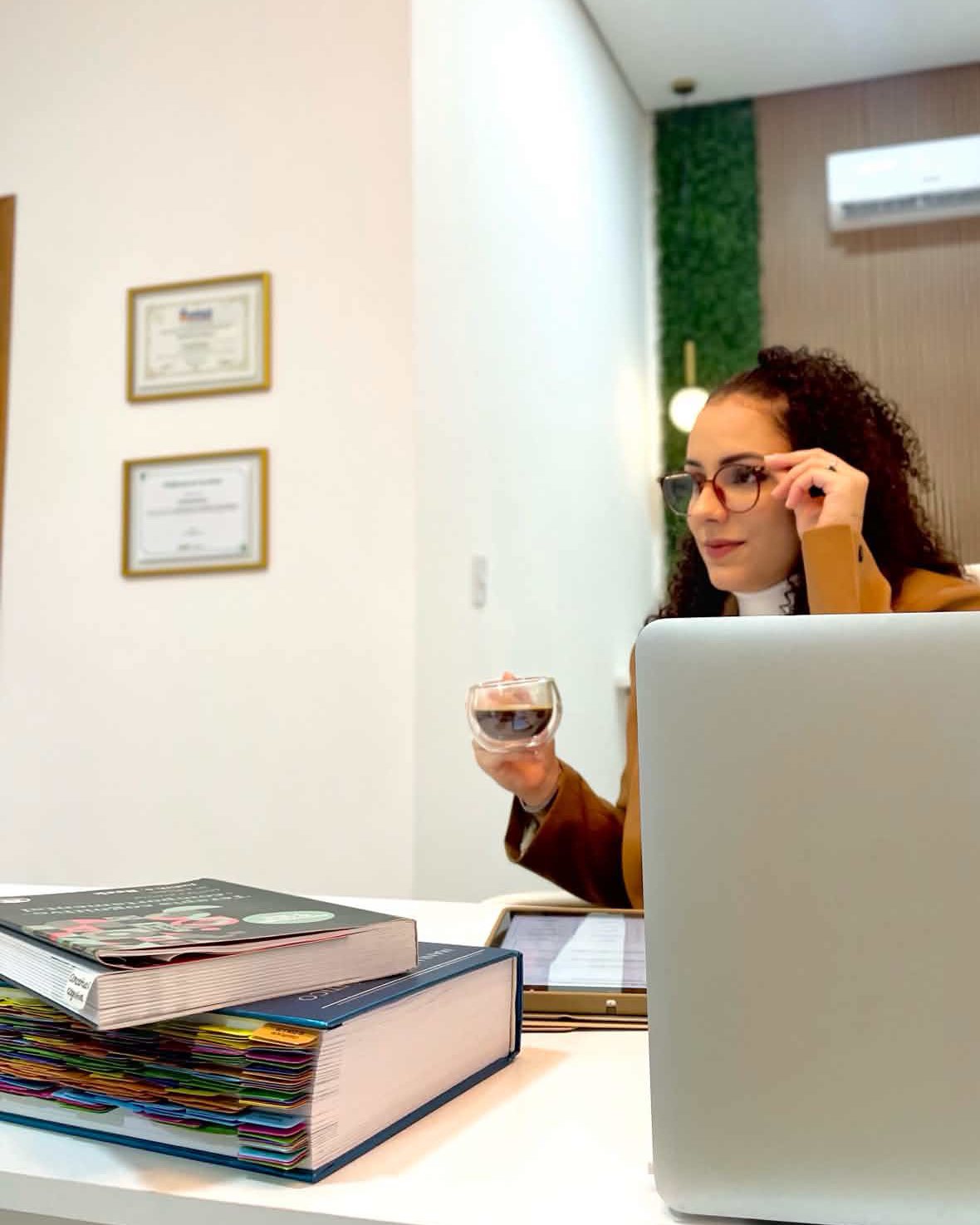 Bruna Ribeiro em seu consultório, ajustando os óculos, com um café na mão e um notebook à frente