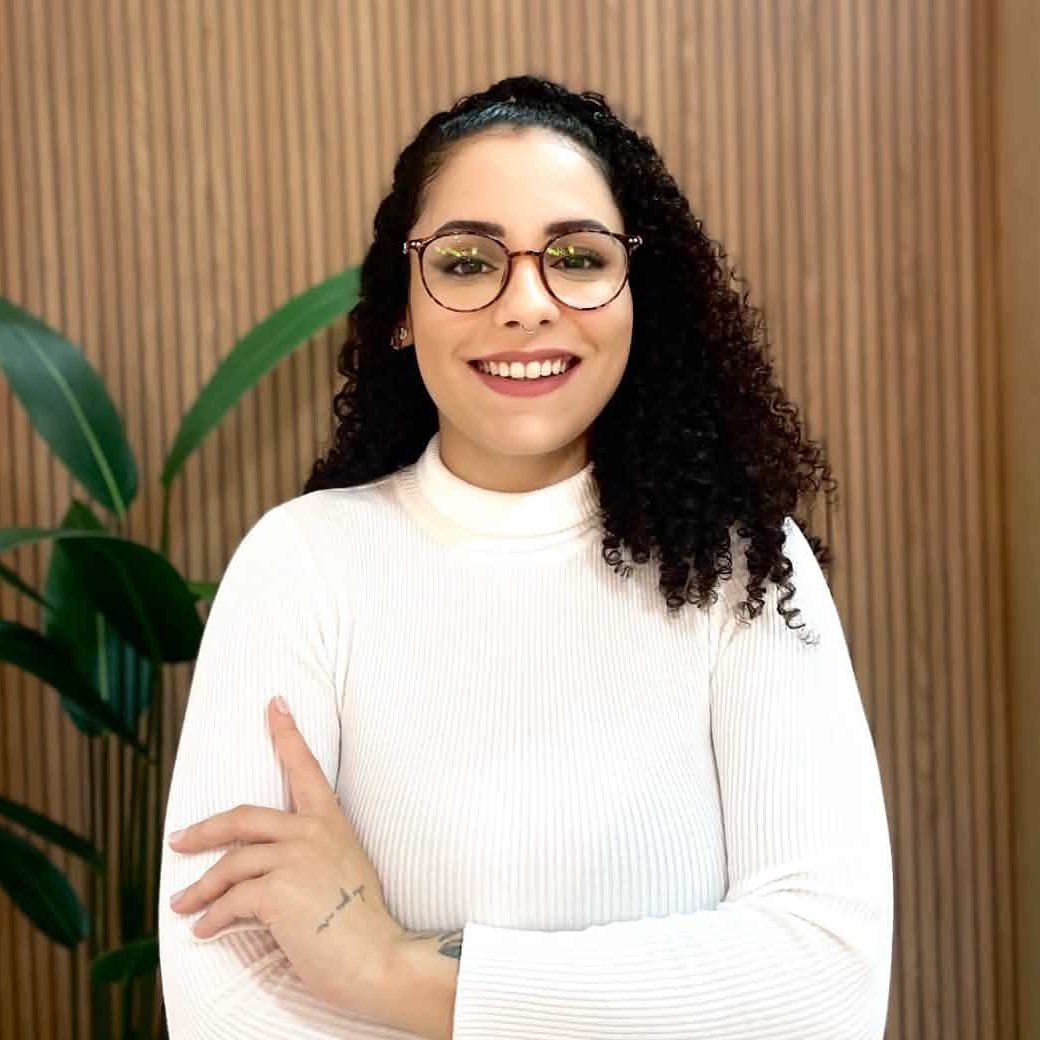 Bruna Ribeiro, psicóloga, sorrindo, de blusa branca e óculos, diante de um painel de madeira com uma planta ao lado