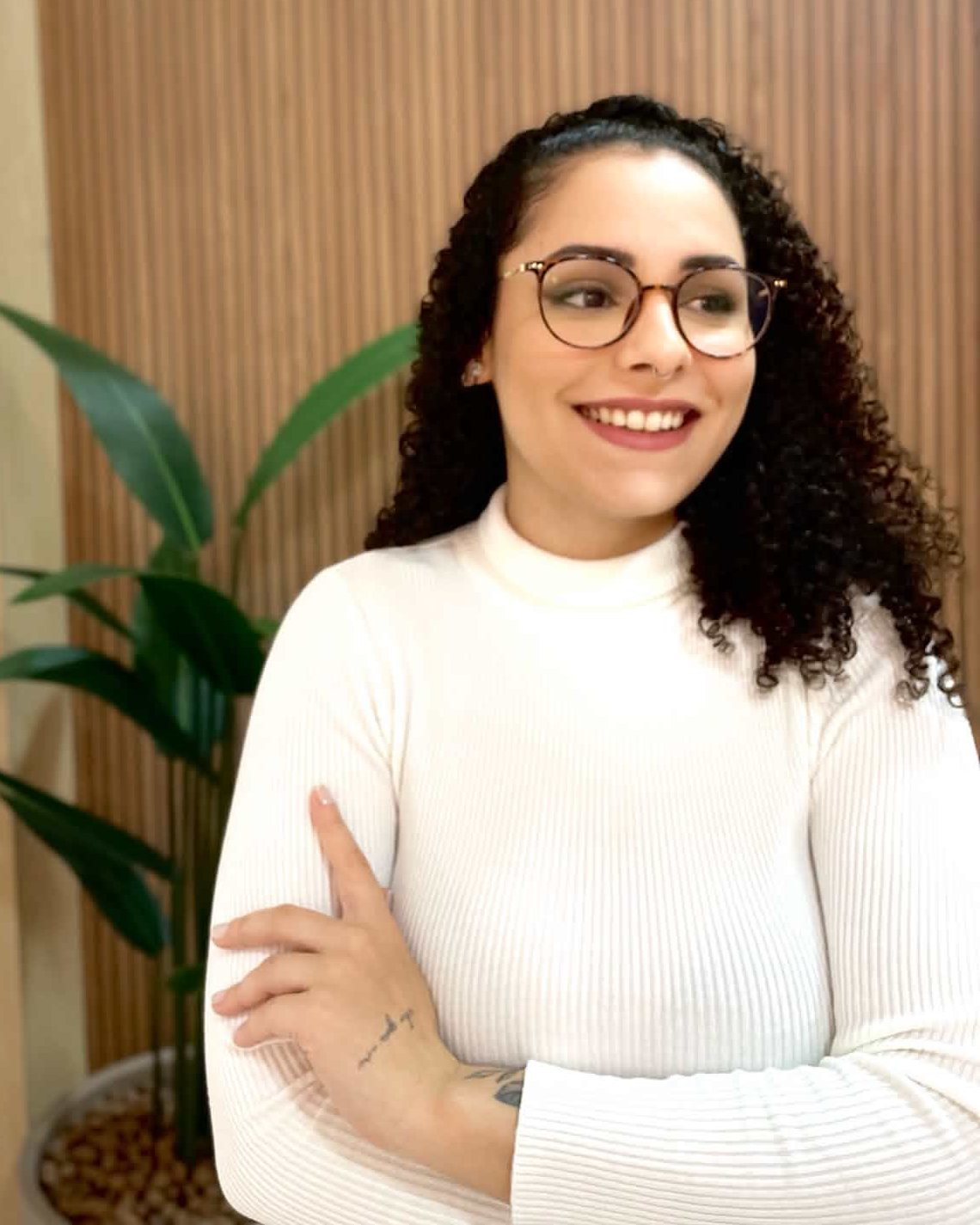 Bruna Ribeiro sorrindo, de braços cruzados, blusa branca e óculos, diante de um painel de madeira